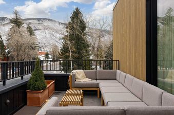 An expansive outdoor terrace featuring a grey L-shaped sofa with wooden accents and a small wooden coffee table, overlooking snow-covered mountains.