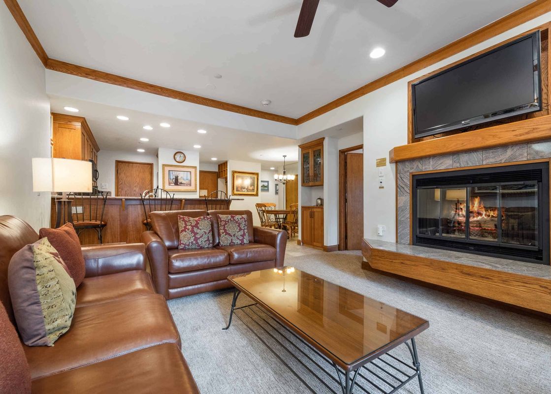 A spacious and inviting living room at The Charter E0405, featuring leather sofas, a lit fireplace, and an open-concept view to the kitchen and dining area.
