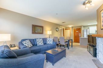 The living area features a comfortable blue sofa and armchair, a coffee table, and a large dining table with chairs, all set against neutral-toned walls.