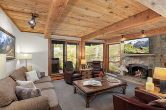 The inviting living room features a comfortable brown sofa, a wood-paneled ceiling with exposed beams, a flat-screen TV above a stone fireplace, a rustic wooden coffee table, and large windows with scenic views.