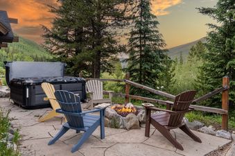 The Royal Retreat features a hot tub, a fire pit area with Adirondack chairs, and stunning views of the surrounding mountains and trees under a colorful sunset sky.