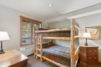 A cozy bunk room in Morning Eagle 304 with a log-style bunk bed, a wooden dresser with a lamp, and a window with blinds overlooking an outdoor view.