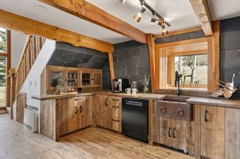 A rustic-style kitchen with natural wood cabinets, slate-accented walls, and an outdoor view through a wooden window frame.