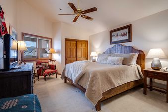A cozy bedroom in Chateau Chamonix CX232, featuring a queen-sized bed with a rustic wooden headboard, soft bedding, and faux fur accents. Two elegant lamps sit on matching nightstands, while a vibrant armchair and a dark wooden dresser complete the inviti