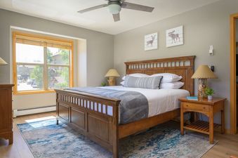 Cozy bedroom in Sawmill Station 217 with a wooden queen bed, rustic bedside lamps, and a window showing the outdoor view.