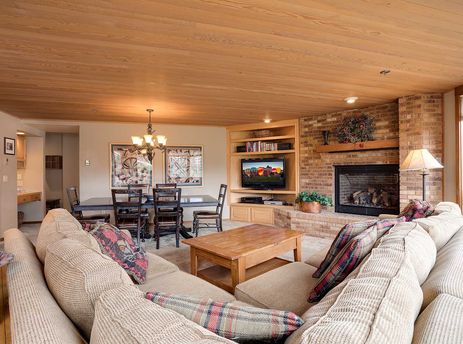 A cozy living room at Chateau Chamonix CX124 features a large sectional sofa, a brick fireplace with a festive wreath, a television, and a dining area with a dark wood table under a chandelier, all beneath a wood-paneled ceiling.