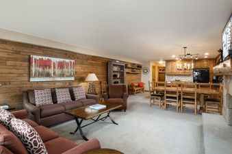 A spacious open-concept living area and dining space at Montaneros 312 featuring warm wood-paneled walls, comfortable brown sofas, a wooden dining table, and a well-appointed kitchen, all under soft lighting.