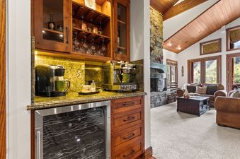 A built-in wet bar with a wine cooler, coffee machines, and glass-fronted cabinets. In the background, a living room with a stone fireplace and seating is visible.