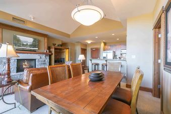 A dining room with a large wooden table and chairs. There is a fireplace in the living room and a kitchen in the background. The room is decorated in a rustic style.