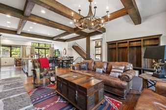 A spacious great room featuring a brown leather sofa, wooden beams, a large chandelier, and a colorful rug, with a kitchen and dining area in the background.