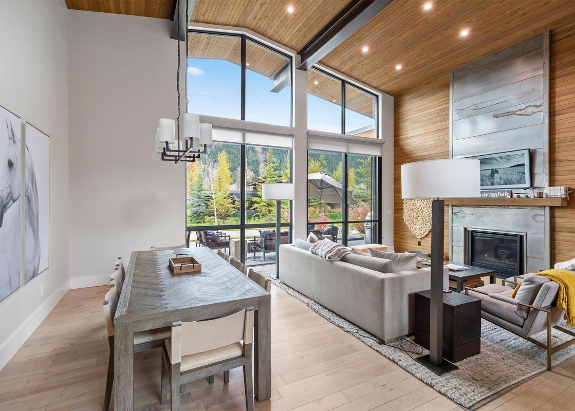 A modern open-plan living and dining area with a long dining table, light-colored sofa, two armchairs, a modern fireplace, and large windows offering views of the exterior natural landscape.