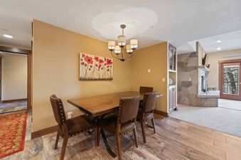 A dining room at The Charter C1405 features a rustic wooden table, four brown leather chairs, and a chandelier, with a red poppy painting on the wall and a stone fireplace in the background.