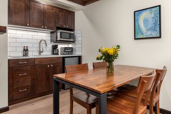 A modern kitchenette with dark wood cabinets, white subway tile, and stainless steel appliances is paired with a wooden dining table and chairs, accented by a vase of yellow flowers and abstract art.