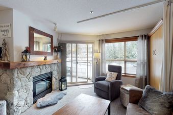 The inviting living area of Aspens on Blackcomb Unit 234 features a stone fireplace, comfortable seating, and a snowy outdoor view.