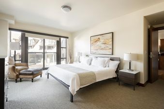 A modern bedroom with a large window looking out onto buildings, featuring a neatly made bed, two nightstands with lamps, a brown armchair with an ottoman, and an abstract painting above the bed.