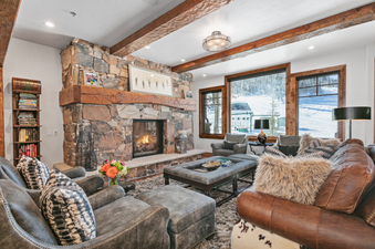 The spacious living room of Grand Lodge 3 Bedroom Deluxe 304 features a cozy fireplace, comfortable leather furniture, and a breathtaking view of the snowy mountain slopes outside. The room has a contemporary design and is well-lit by natural sunlight. Th