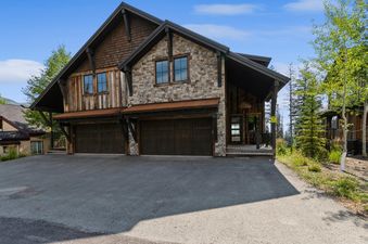 The exterior of a large, rustic mountain home featuring stone and wood siding, a double garage, and a welcoming entrance with natural landscaping under a blue sky.