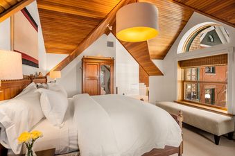 A cozy bedroom with a large bed featuring white linens, a wooden vaulted ceiling, and an arched window offering a view of the outside.