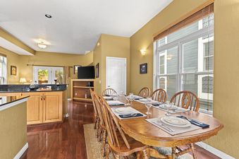 A spacious dining area featuring a large wooden table set for a meal, surrounded by wooden chairs, with warm golden walls, hardwood floors, and natural light from a large window.