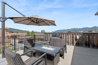A spacious outdoor deck with a fire pit table, woven chairs, a patio umbrella, and a BBQ grill, offering mountain and town views.
