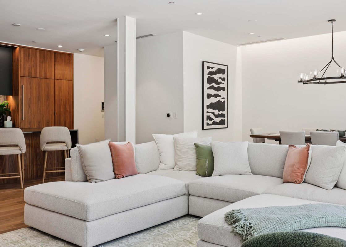 An expansive open-concept living area featuring a large, light-colored sectional sofa with decorative pillows, facing a modern kitchen with dark and wood cabinetry, and a dining area with a chandelier in the background.