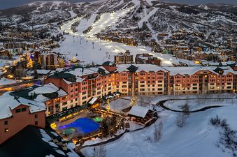 An aerial inverted night view of the Steamboat Grand Deluxe King resort complex, featuring snow-covered buildings, illuminated ski slopes, and a vibrant blue outdoor pool area.