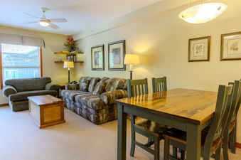 The living area within Aspens on Blackcomb Unit 438 features a comfortable sofa and armchair, a coffee table, and a dining area with a wooden table and chairs, all illuminated by a ceiling fan with light and lamps.