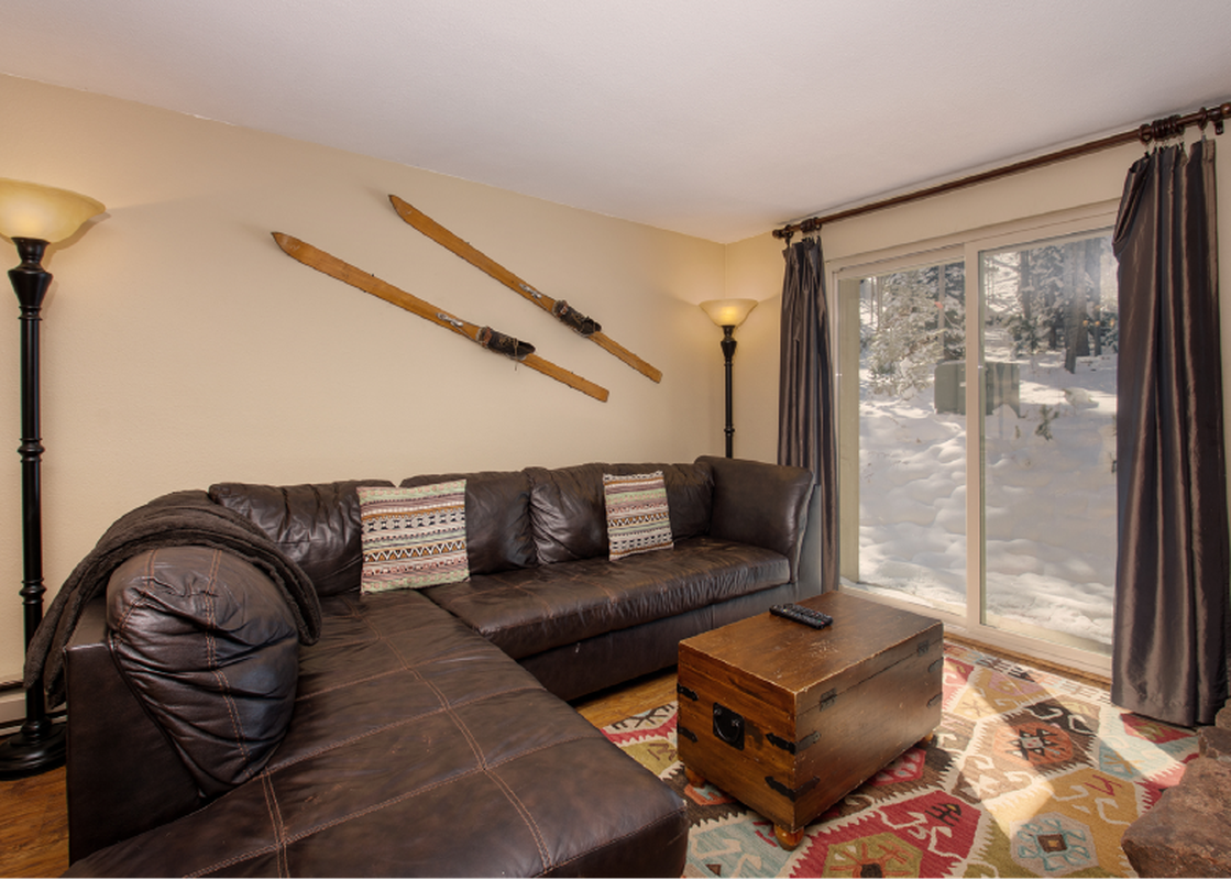 A spacious living area featuring a large brown sectional sofa, a wooden coffee table, and a colorful patterned rug. The room is illuminated by two floor lamps, and vintage wooden skis adorn one wall, while a glass sliding door reveals a snowy outdoor scen