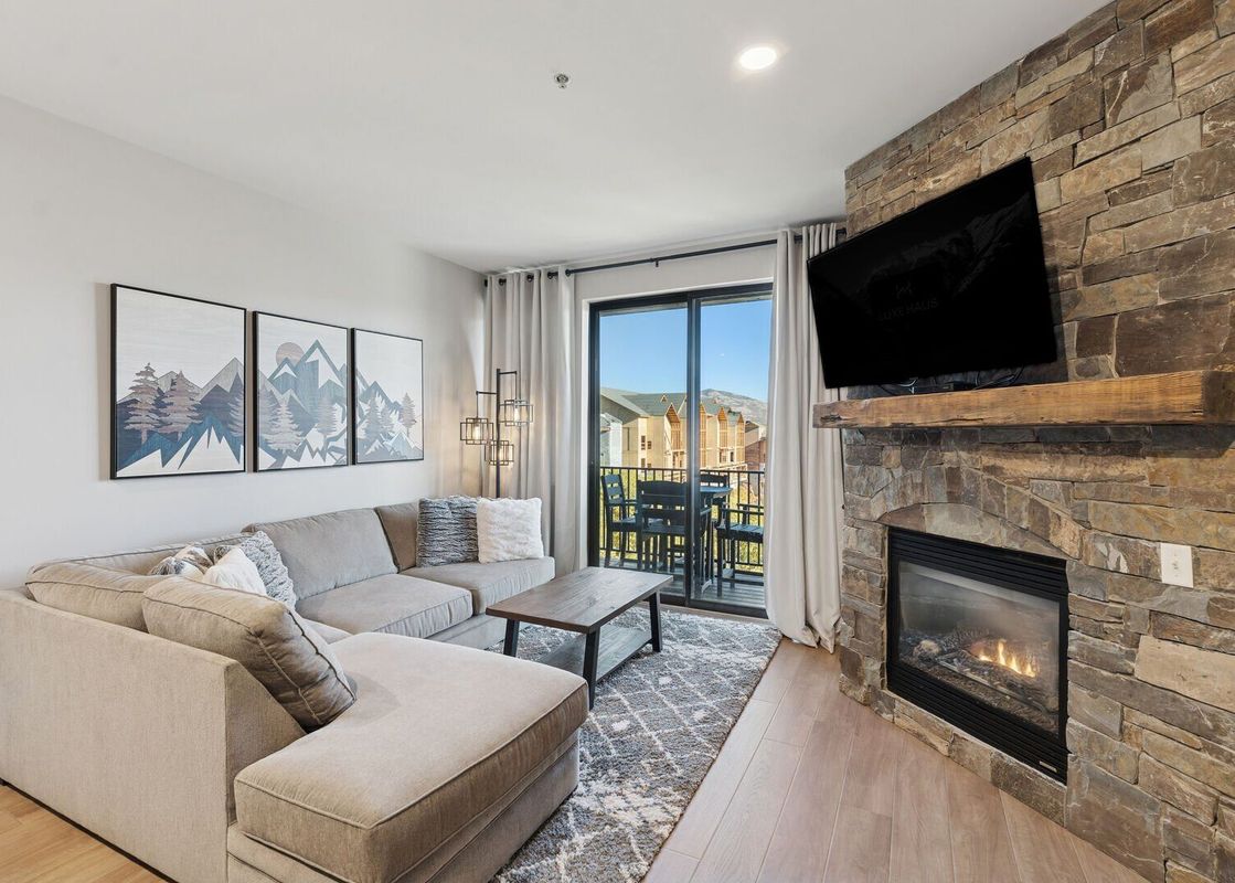 A cozy living room in The Mason 3026 featuring an L-shaped sectional sofa, a stone fireplace with a TV, and a balcony with mountain views.