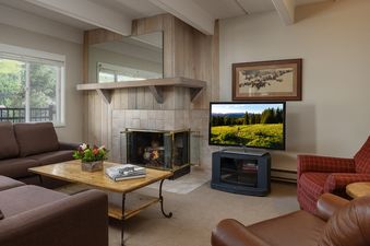 The living room at Vantage Point 313 features a comfortable brown sectional, a fireplace, a TV, and a red armchair, with a large window showcasing mountain views.