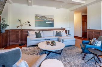 The living room of Victoria Condominiums 2 Bedroom features a neutral-toned sectional sofa, a round coffee table, and blue and grey armchairs, all set on a patterned area rug with wood wainscoting and a fireplace.