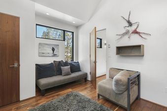 A modern room features a grey daybed with pillows, a beige armchair, wooden flooring, and contemporary art, all bathed in natural light from a large window overlooking trees.