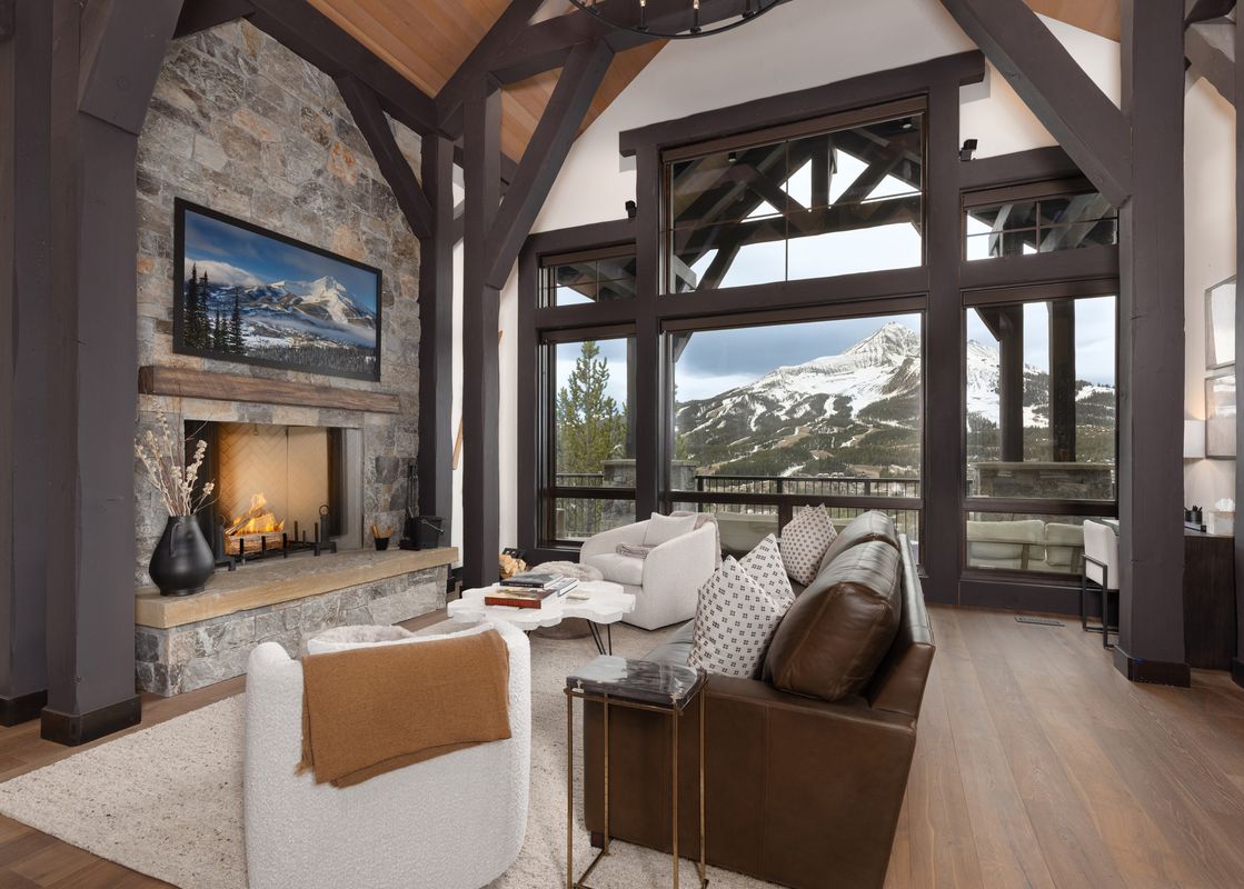 A cozy living room with a stone fireplace, large windows overlooking a snowy mountain, and comfortable seating.