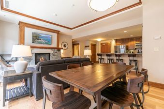 A modern living room with a fireplace, a large flat screen TV, a wood dining table, and a fully equipped kitchen.
