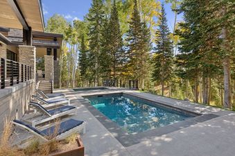An outdoor pool and hot tub area at All In at The Colony, surrounded by lounge chairs and backed by a dense forest with autumn foliage.