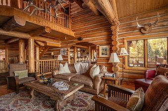 A cozy, rustic lounge in Huckleberry Hideaway featuring log walls, comfortable seating, and a large window overlooking a forest.