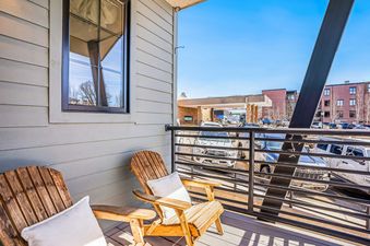 A wooden balcony with two chairs and a view of a parking lot and a building in the distance.