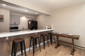 A modern kitchen and dining area in Meadow Ridge 18 Unit 11, featuring gray cabinets, white subway tile backsplash, and a black refrigerator, complemented by a white granite countertop with four wooden bar stools and a long wooden dining table set for two