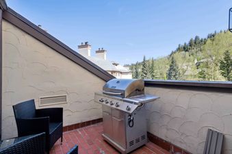 An outdoor patio at The Charter L222 with a stainless steel BBQ grill, black wicker chairs, and a red brick floor, set against a backdrop of green trees and mountains under a blue sky.