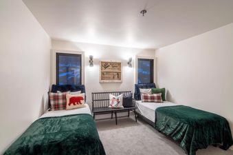A cozy bedroom in Le Chamonix Unit A features two twin beds with dark green blankets, plaid and bear-themed pillows, and a ski-themed sign on the wall between two windows.