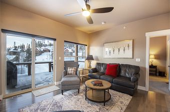 A spacious and inviting living room at Rendezvous 47 Lookout Point, featuring a comfortable leather sofa, stylish armchair, and a circular coffee table, all bathed in natural light from large windows and a sliding glass door leading to a snowy balcony.