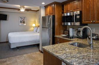 A comfortable studio unit at Breckenridge Studio Beaver Run Resort featuring a compact kitchen with granite countertops, stainless steel appliances, and a bedroom area with a cozy bed, bedside lamp, and wall-mounted TV.