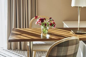 A wooden table with two chairs and a vase of pink and red flowers in a light and airy room