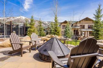 An outdoor patio at EagleRidge Townhomes 1461 features a black fire pit surrounded by four Adirondack chairs, with snow-capped mountains and townhomes in the background under a blue sky.