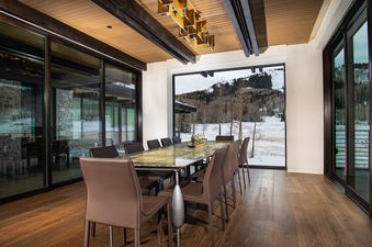 The dining room at White Pine Canyon features a long table with dark brown leather chairs. The table has a stone countertop with a marble-like finish and features a large panoramic window with mountain views and the snow-covered landscape outside.