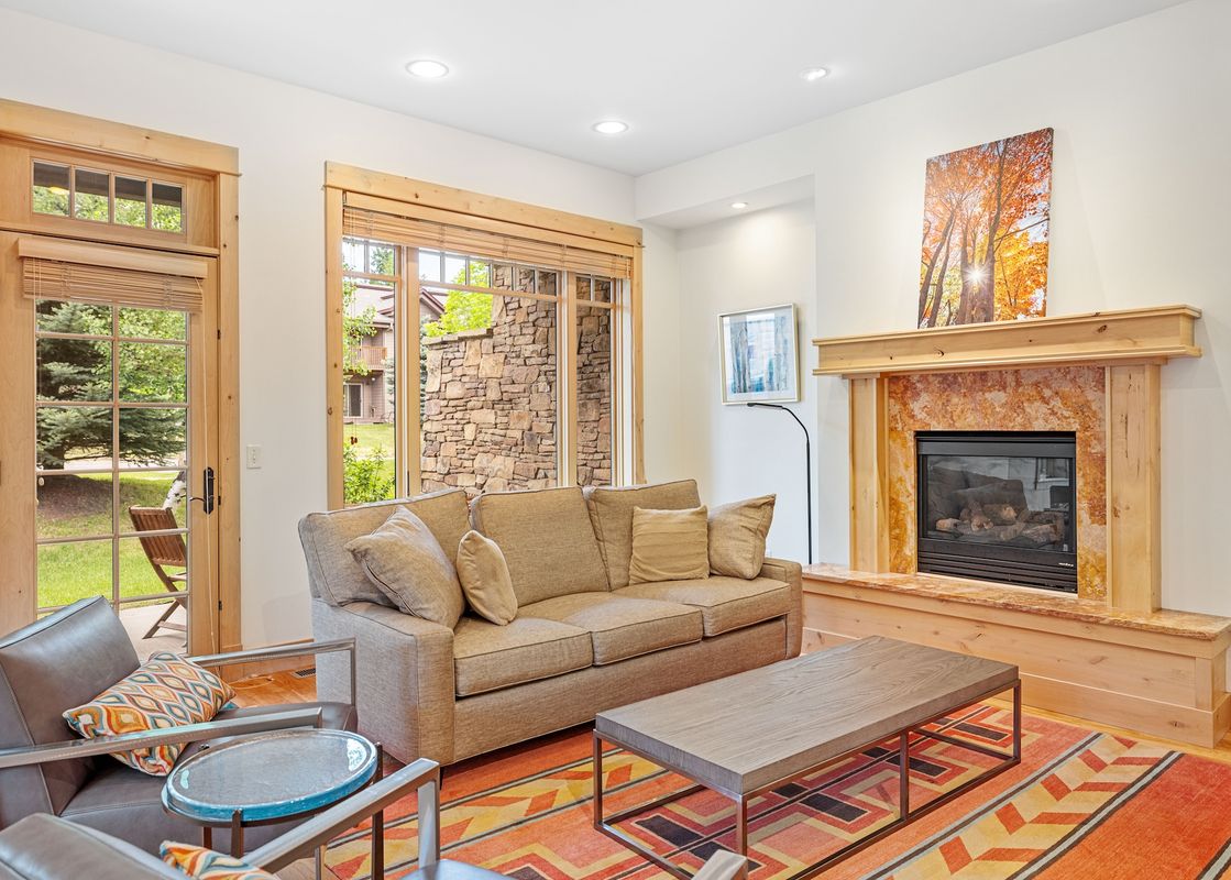 A cozy living room featuring a fireplace, a comfortable sofa, armchairs, and a vibrant geometric rug, with a large window and glass-paneled door offering views of the outdoors.