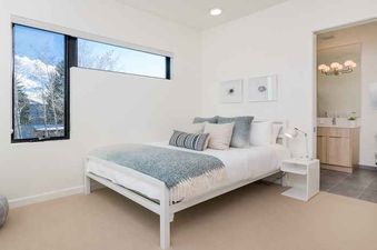 A bedroom with a white bed and a blue blanket, a window with a view of the mountains, and a bathroom in the background.