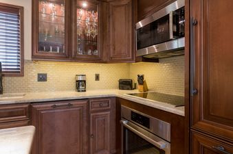 A modern kitchen with dark wood cabinets, light-colored tile backsplash, stainless steel appliances, and glass-front upper cabinets displaying glassware.