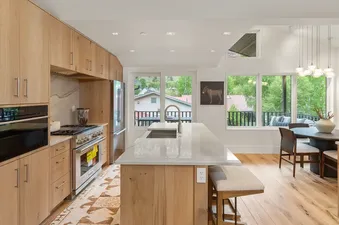 A modern kitchen with light wood cabinetry, stainless steel appliances, a kitchen island with a sink, and a dining area with a round table and pendant lights, all overlooking a green exterior view.