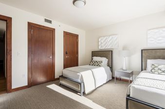Two twin beds with white linens and patterned pillows in a light and airy bedroom with wood floors and large windows.